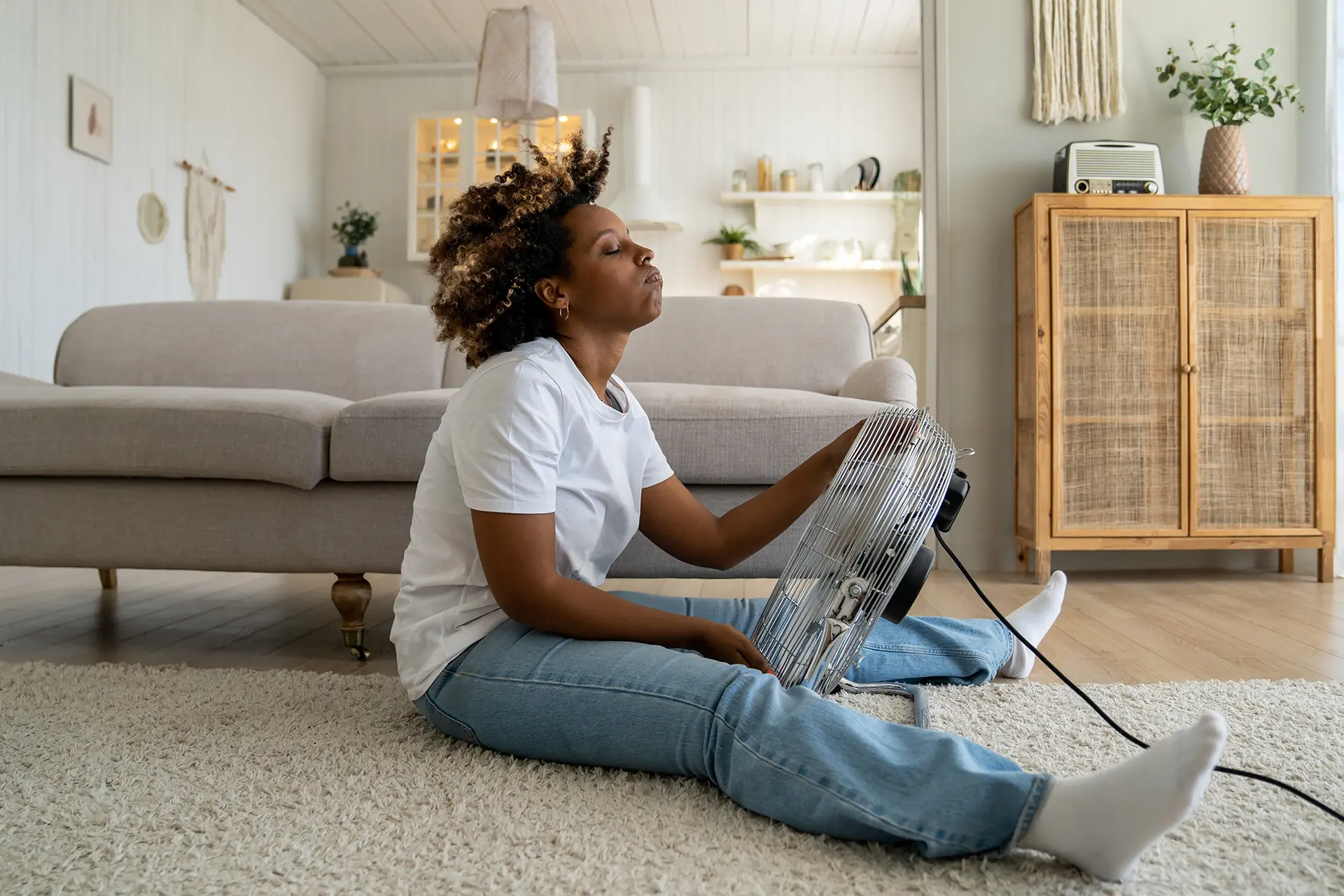 Tired overheated black woman cooling herself with electric fan at home, suffering from heat while sitting on floor in living room without conditioner, keeping cool indoors during summer heatwave