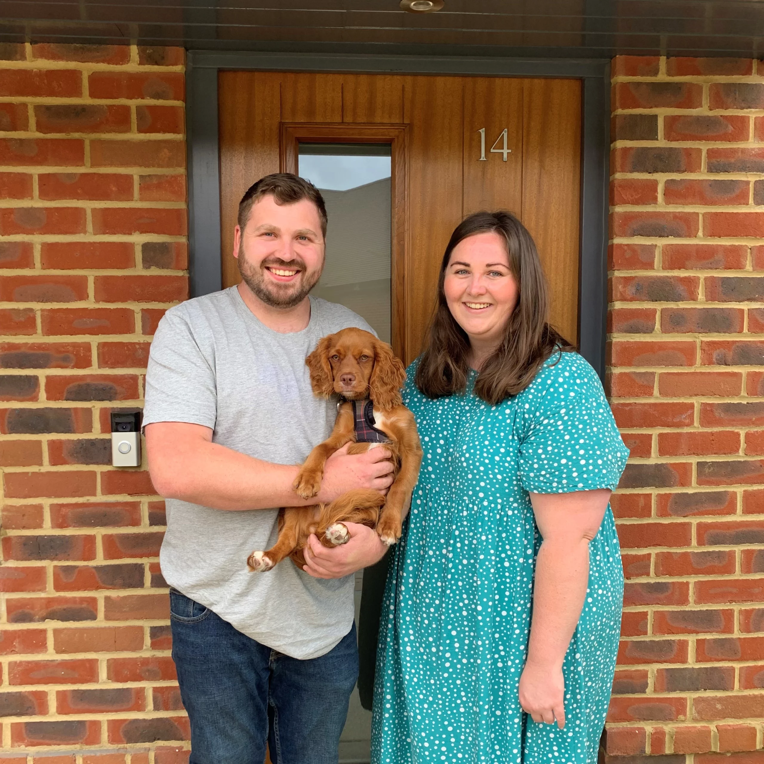 A couple stand in front of the front door of a new home holding a dog